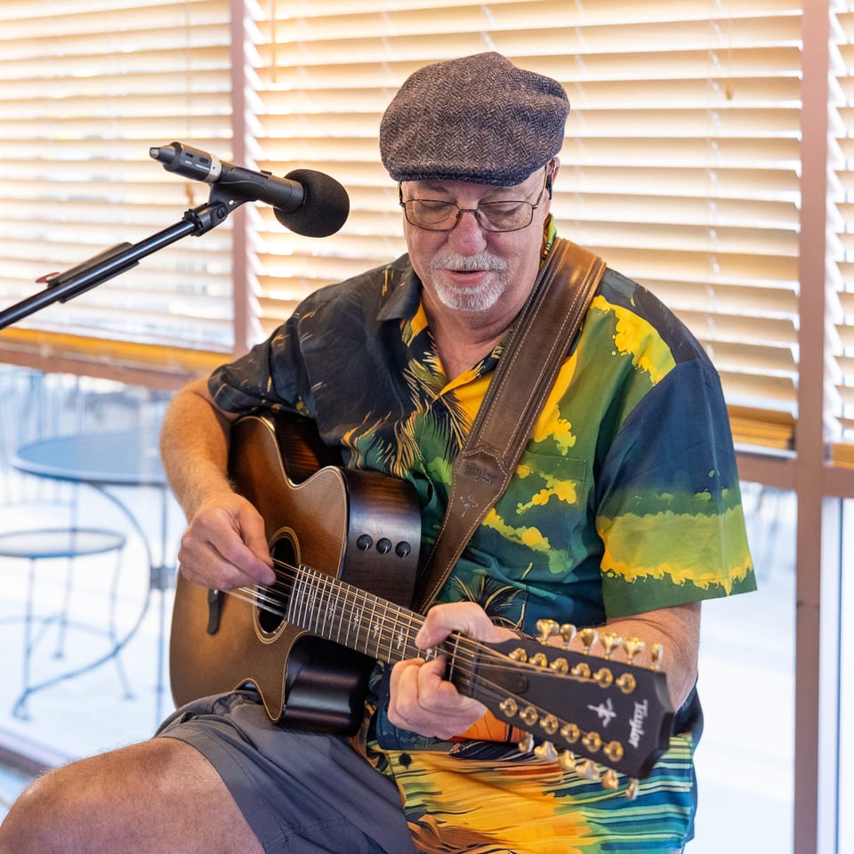 Musician in a flat cap and colorful shirt plays acoustic guitar beside a microphone indoors.