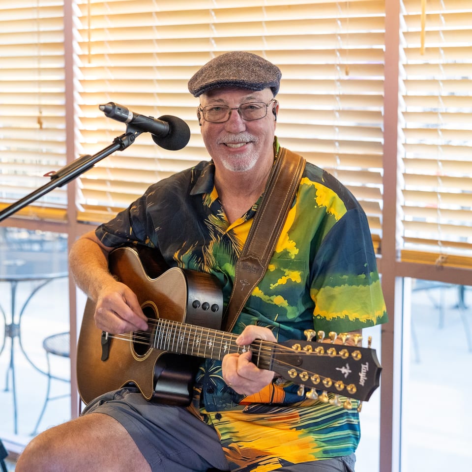 Musician in a colorful shirt and flat cap plays acoustic guitar beside a microphone in a bright restaurant.