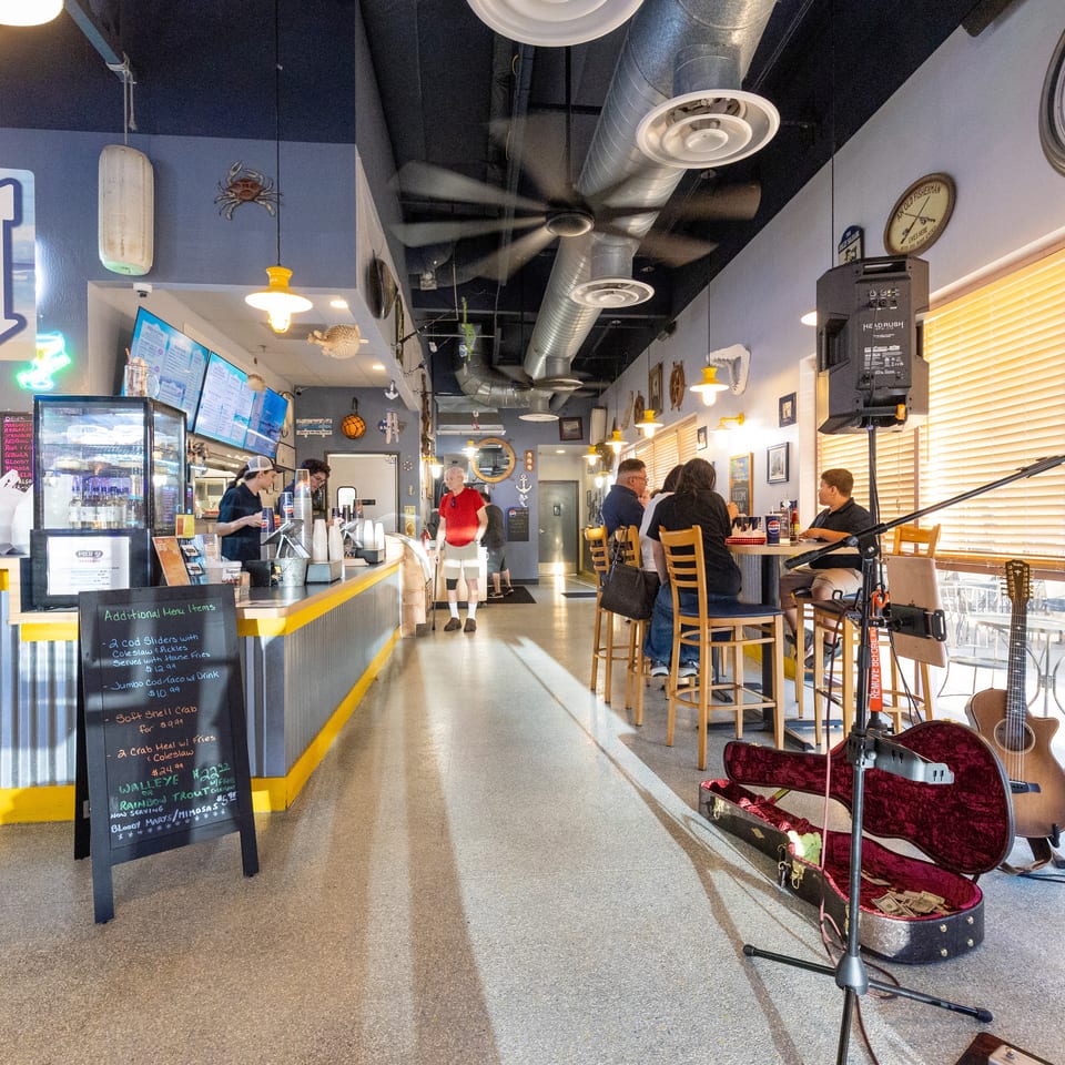 Bright nautical-themed seafood restaurant interior with counter service, diners, menu boards, maritime décor, and a musician’s guitar setup.