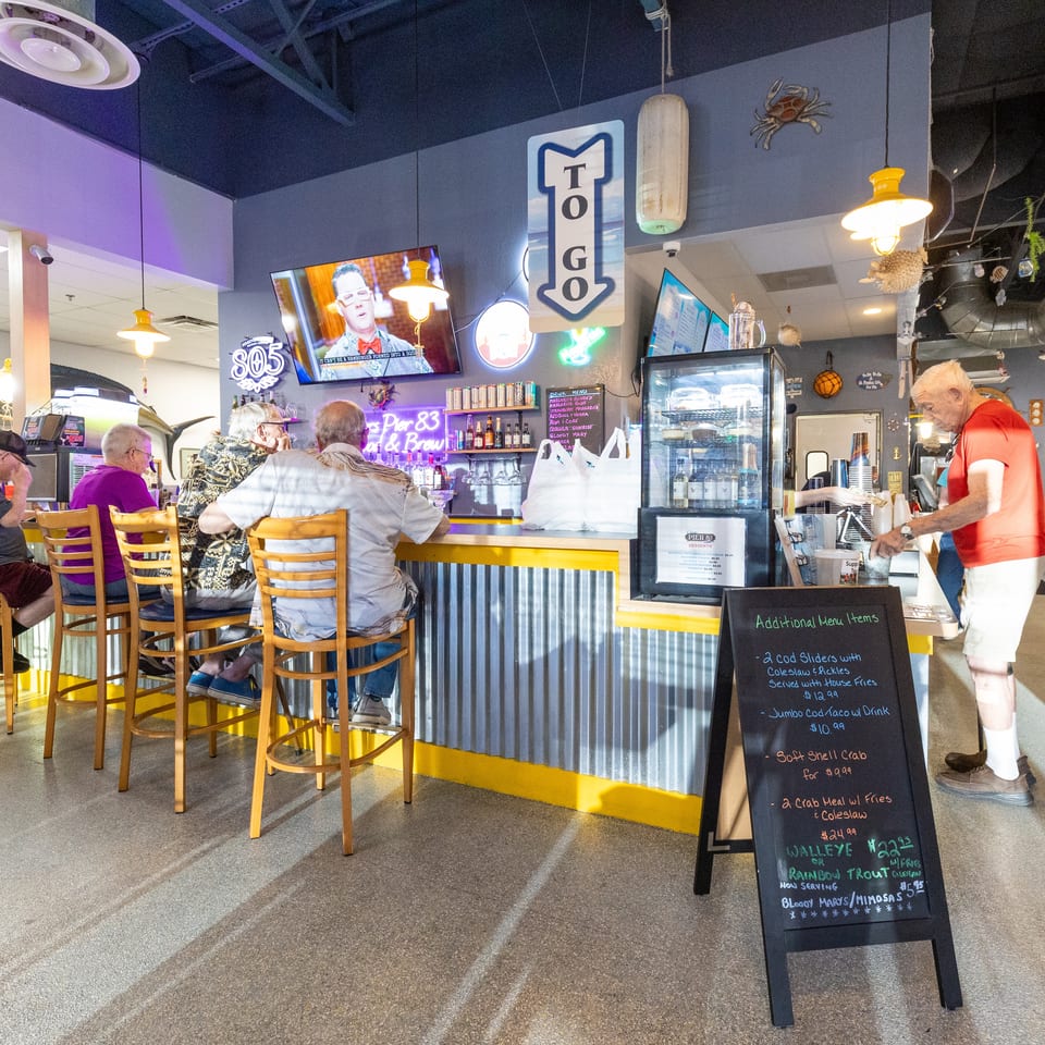 Diners sit at a colorful seafood restaurant counter while staff serve customers beneath nautical décor and televisions.