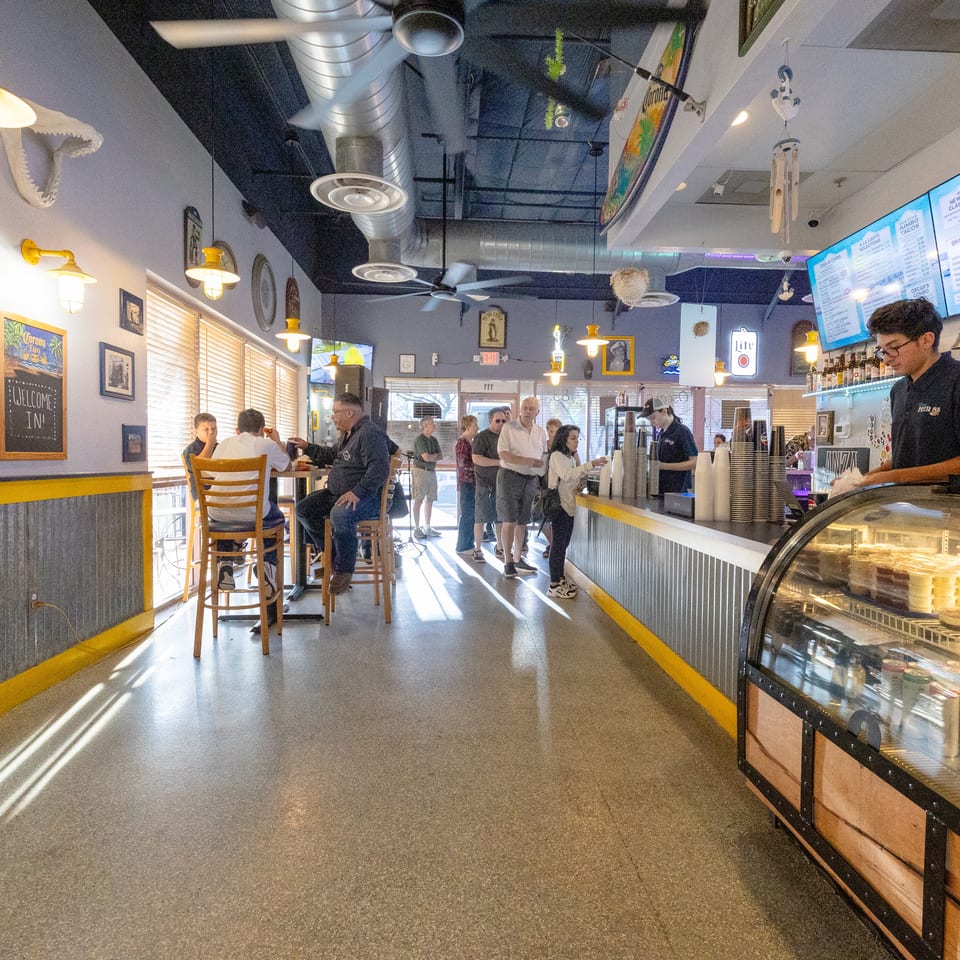 Busy casual seafood restaurant interior with diners, counter service, digital menus, nautical decor, and a refrigerated display case.