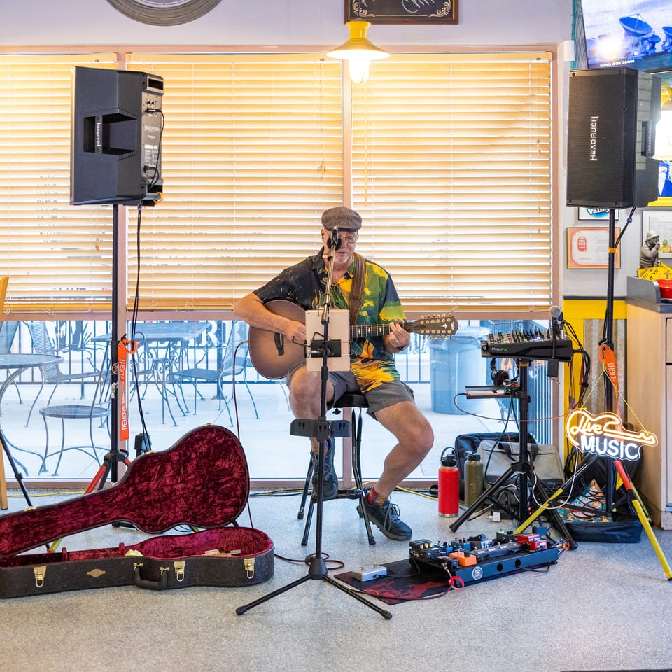Musician in a Hawaiian shirt performs with an acoustic guitar amid speakers, microphones, and music equipment inside a restaurant.