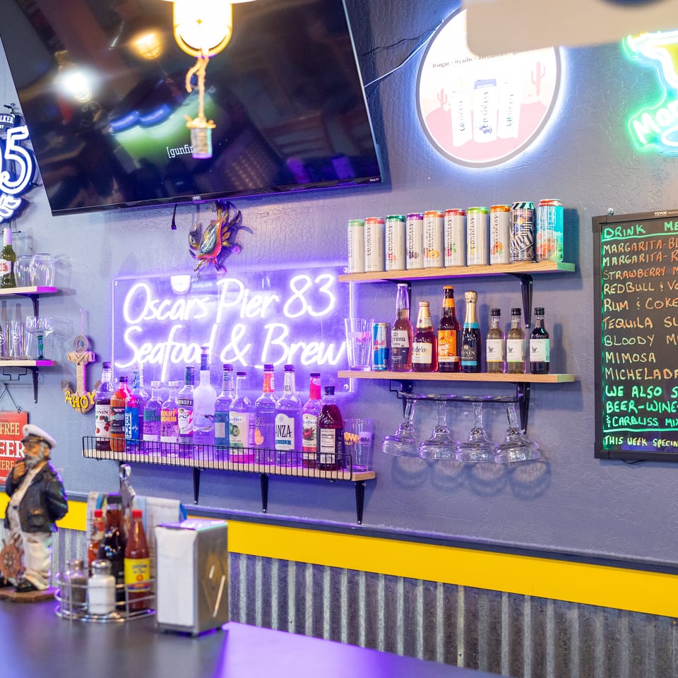 Colorfully lit restaurant bar with liquor bottles, beer, glassware, neon signs, television, and a handwritten drink menu.