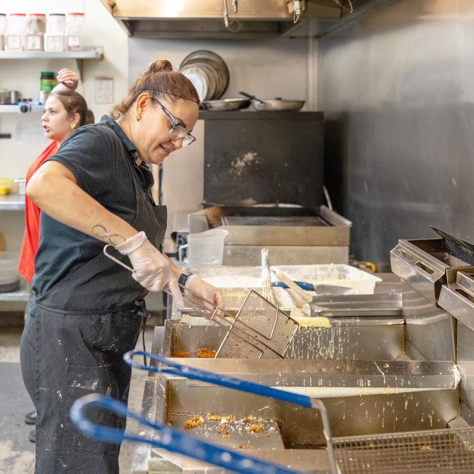 Restaurant kitchen worker in gloves operates a fryer while another employee works behind her.