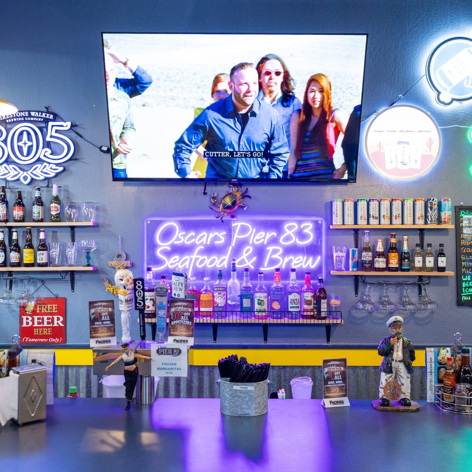 Colorful bar at Oscars Pier 83 Seafood & Brew, featuring beer and liquor shelves, neon signs, a television, and drink menu.