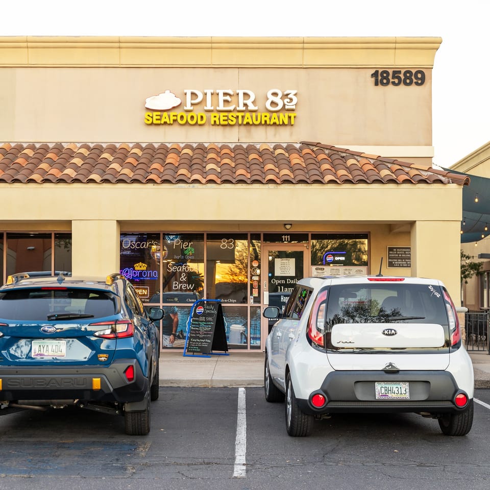 Pier 83 Seafood Restaurant storefront with tiled roof, glowing signage, outdoor seating area, and parked cars.