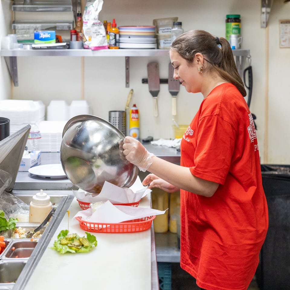 Restaurant worker in a red shirt pours prepared ingredients from a metal bowl into a lined basket at a food preparation counter.