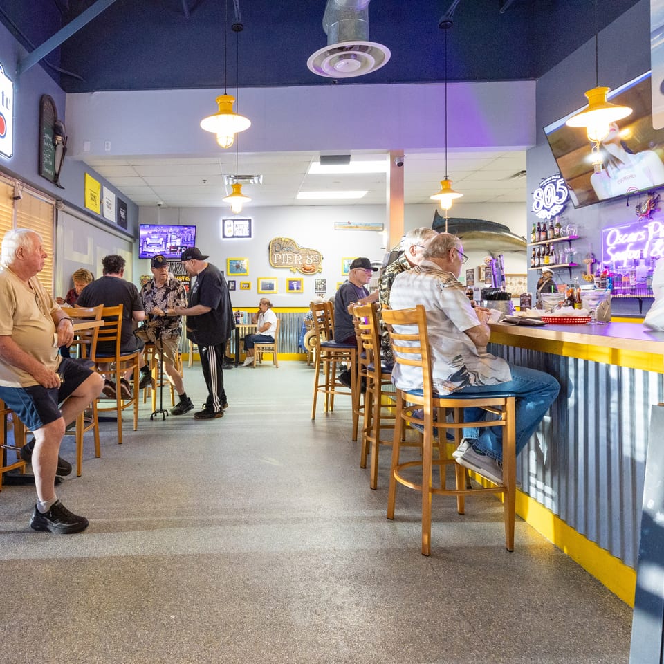 Lively casual restaurant interior with diners seated at tables, a bar, neon signs, televisions, and nautical decor.