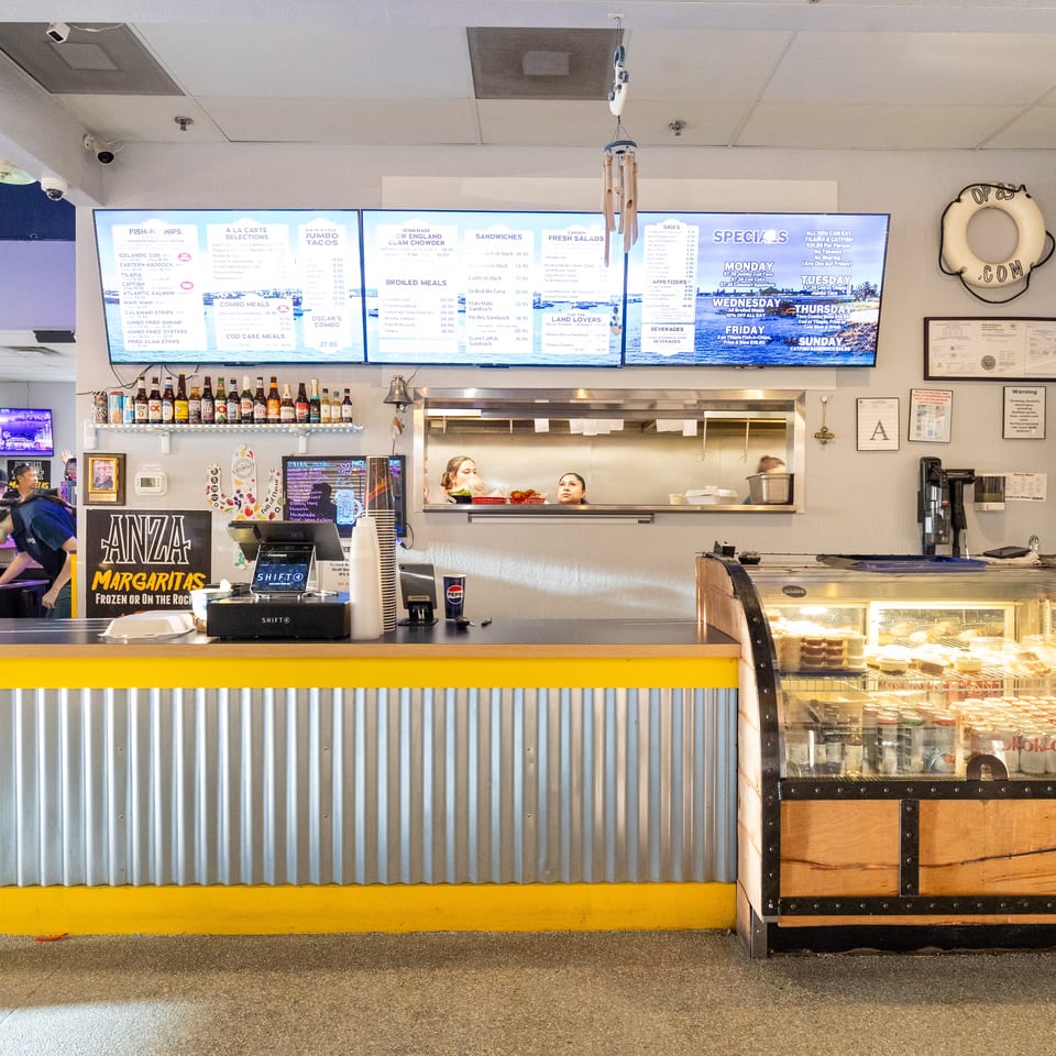 Seafood restaurant counter with illuminated menus, staff serving customers, bottled drinks, and a dessert display case.