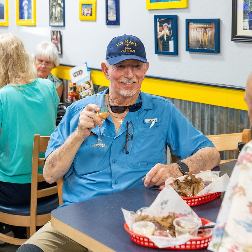 Man wearing a U.S. Navy veteran cap raises a drink at a lively restaurant table surrounded by diners and framed photographs.