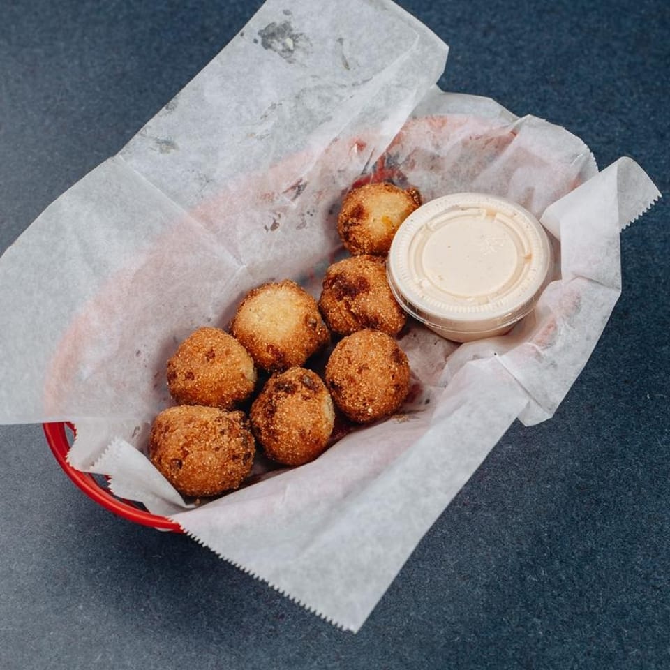 Basket of golden fried hush puppies served with a cup of creamy dipping sauce.